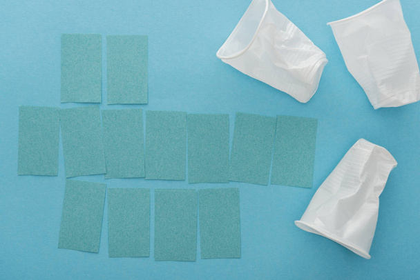 top view of white plastic cups and empty sticky notes on blue background  - Foto, Bild