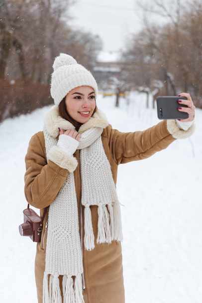 Young woman in hat and scarf walking outdoors in winter snow using mobile phone. - Fotoğraf, Görsel