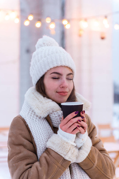 Young woman in hat and scarf walking outdoors in winter snow drinking coffee. - Fotó, kép