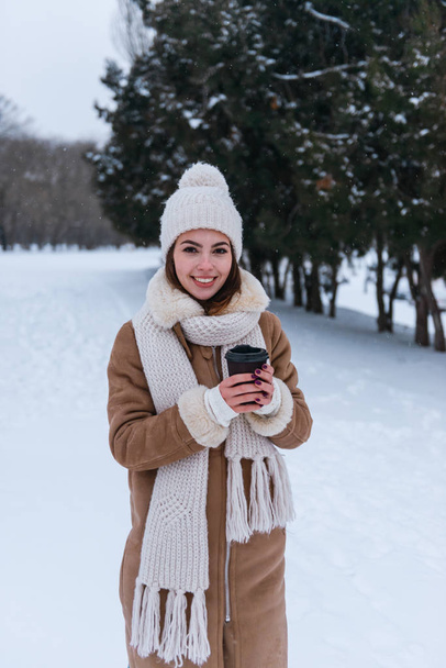 Young woman in hat and scarf walking outdoors in winter snow drinking coffee. - Foto, imagen