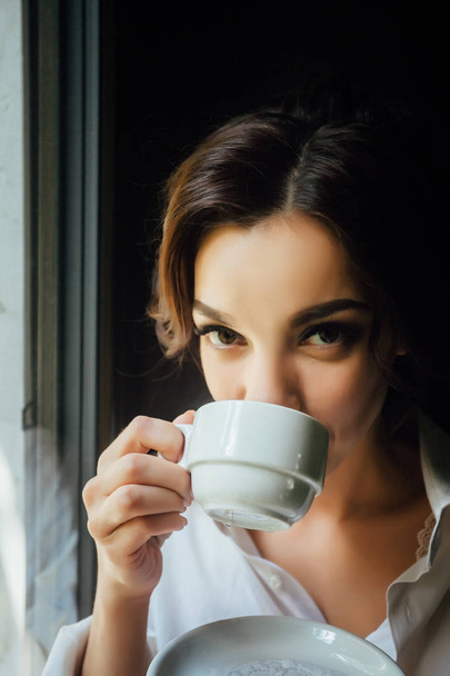 Een mooi meisje zit op de vensterbank in de buurt van een groot raam en haar's ochtends koffie drinken - Foto, afbeelding