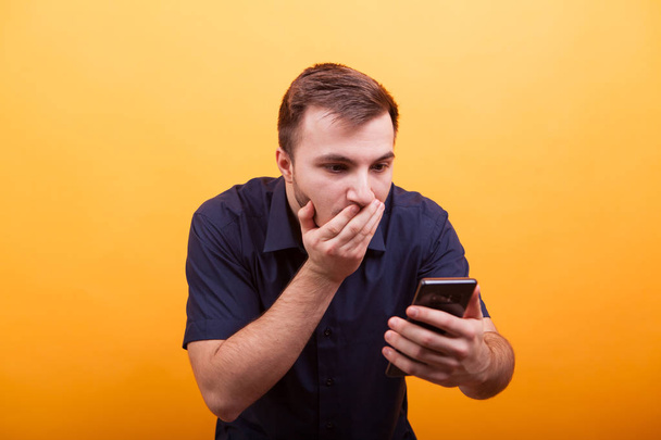 Portrait of shocked young man looking at mobile phone isolated on yellow background - Foto, afbeelding