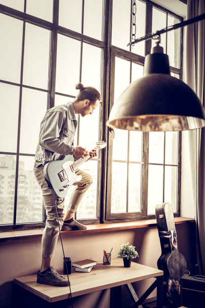 Rock musician standing on table near window and playing the guitar - Foto, imagen