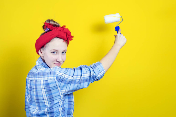 Young beautiful girl in a plaid shirt and a red bandage on his head, paints with a paint roller against the background of a yellow studio. Retro style. - Zdjęcie, obraz