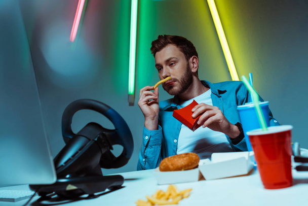 handsome and good-looking man eating french fries and looking at computer monitor  - Foto, Imagen