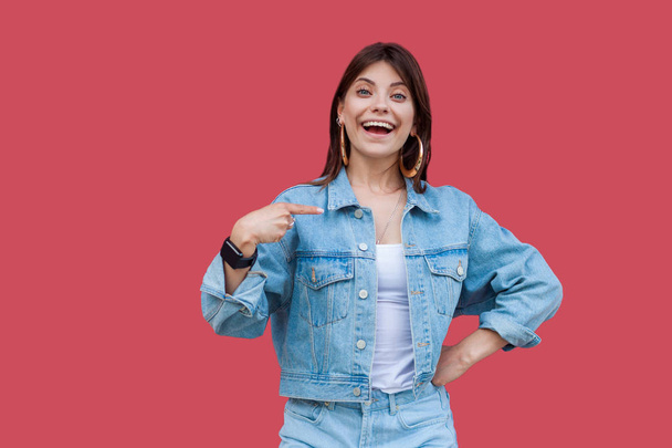 Portrait of surprised beautiful brunette young woman with makeup in denim casual style pointing herself and looking at camera with amazed face on red background - Фото, изображение