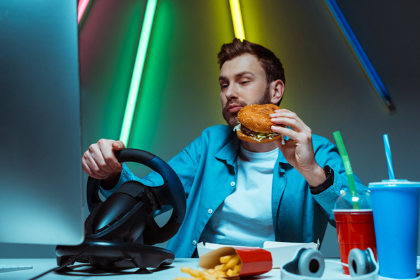 selective focus of handsome cyber sportsman eating burger and playing computer game   - Zdjęcie, obraz