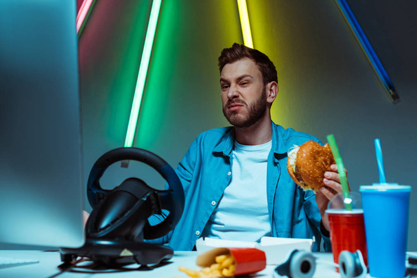 selective focus of handsome man eating burger and looking at computer monitor  - Photo, Image