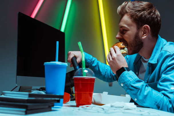 selective focus of handsome and good-looking cyber sportsman eating tasty burger and playing video game  - Фото, изображение
