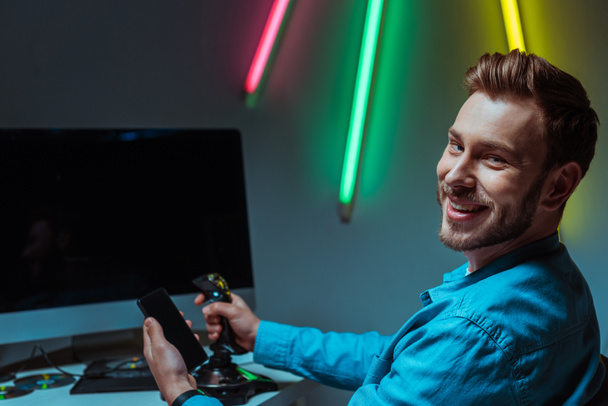 selective focus of smiling and handsome man holding joystick and smartphone  - Fotoğraf, Görsel