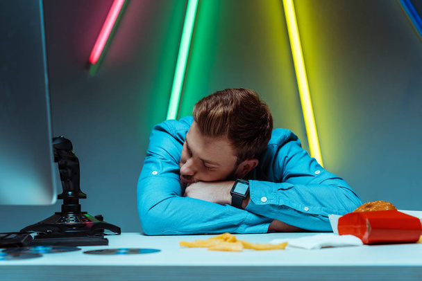  schöner junger erwachsener Mann schläft und ruht auf Tisch mit Steuerknüppel und Fast Food  - Foto, Bild