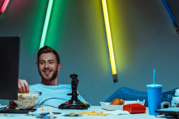 handsome and smiling man looking at computer monitor and eating popcorn  - Photo, Image