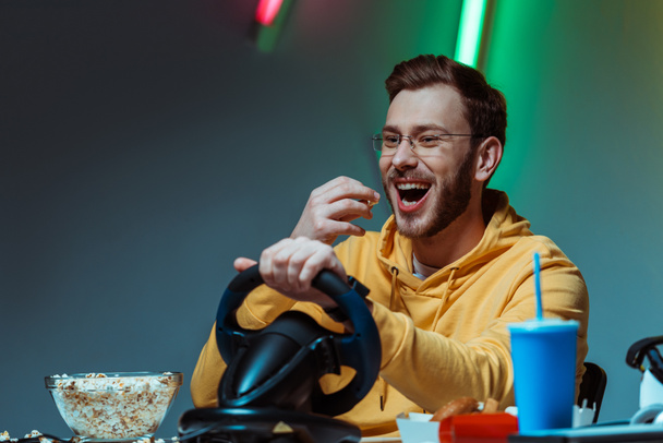 uśmiechnięty i dobrze wyglądający mężczyzna w okularach gra z kierownicy i jedzenia popcorn  - Zdjęcie, obraz