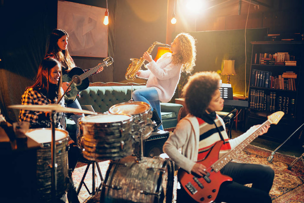 Girls playing jazz music. In foreground one woman playing bass guitar and in background other two playing saxophone and drums. Home studio interior. - Foto, Imagem