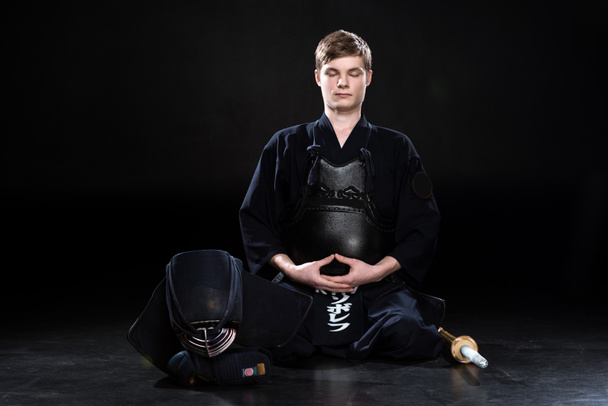 Νεαρός άνδρας στο kendo πανοπλία κάθεται με κλειστά τα μάτια σε μαύρο - Φωτογραφία, εικόνα