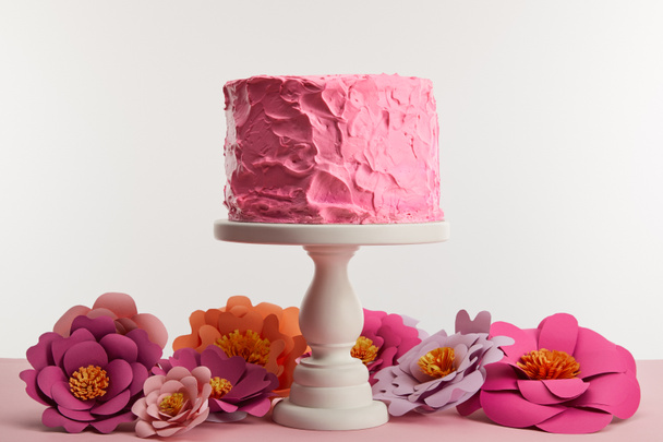 pink birthday cake on cake stand near paper flowers isolated on grey - Foto, Imagen