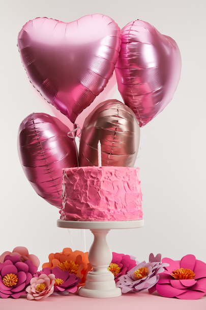 tasty pink birthday cake with candle on cake stand near paper flowers and heart-shaped air balloons on grey - Photo, Image