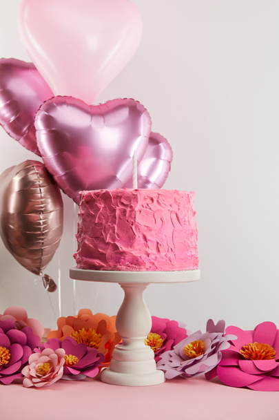 sweet pink birthday cake with candle on cake stand near paper flowers and heart-shaped air balloons on grey - Fotoğraf, Görsel