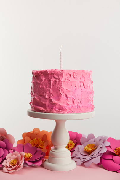 delicious pink birthday cake with candle on cake stand near paper flowers isolated on grey - Photo, Image