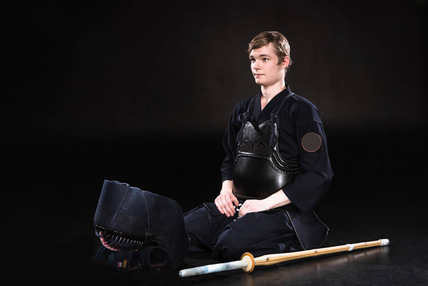 junger Mann in Kendo-Rüstung mit Helm und Bambusschwert sitzt auf schwarz - Foto, Bild