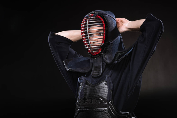 Kendo fighter in armor tying helmet and looking away on black - Fotoğraf, Görsel