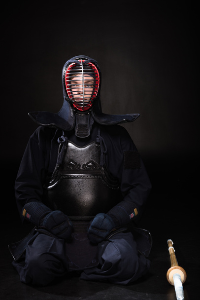 Kendo fighter in helmet with bamboo sword sitting on black - Zdjęcie, obraz