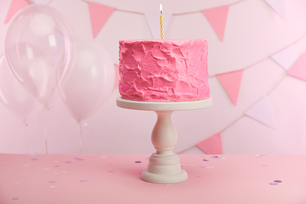 tasty pink birthday cake with burning candle on cake stand near air balloons and decoration - Zdjęcie, obraz