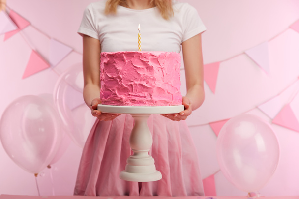 cropped view of woman holding cake stand with sweet pink birthday cake and burning candle near air balloons and decoration - Φωτογραφία, εικόνα