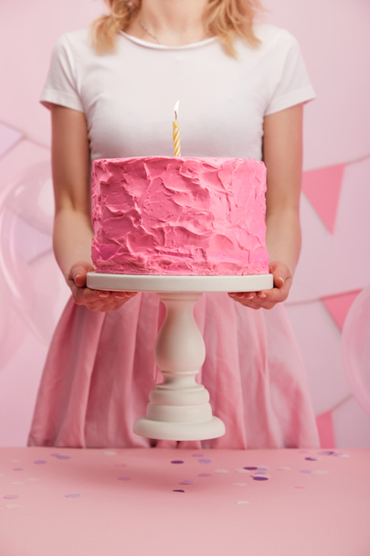 bijgesneden beeld van vrouw met cake stand met zoete roze verjaardagstaart en brandende kaars  - Foto, afbeelding