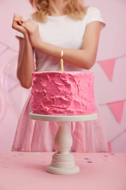 vista recortada de la mujer de pie cerca del soporte de la torta con sabroso pastel de cumpleaños rosa y vela ardiente
 - Foto, imagen