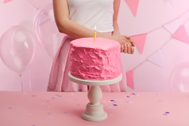 cropped view of woman standing near sweet and tasty pink birthday cake with burning candle on cake stand  - Photo, Image
