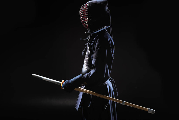 Side view of kendo fighter in traditional helmet holding bamboo sword on black - Zdjęcie, obraz
