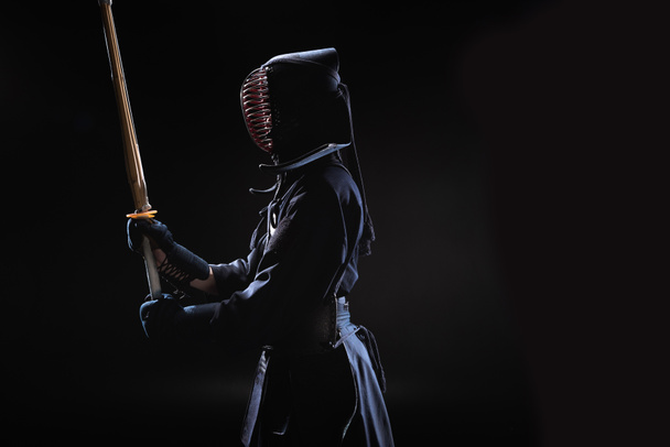 Vista lateral de kendo fighter en casco tradicional sosteniendo espada de bambú sobre negro
 - Foto, imagen