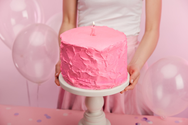 bijgesneden beeld van vrouw houden van taart stand met roze verjaardagscake en branden van de kaars van de nummer één in de buurt van lucht ballonnen - Foto, afbeelding