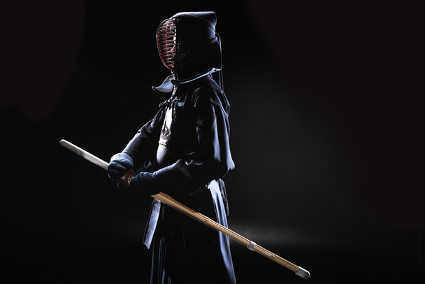 Vista lateral de kendo fighter en casco tradicional sosteniendo espada de bambú sobre negro
 - Foto, Imagen