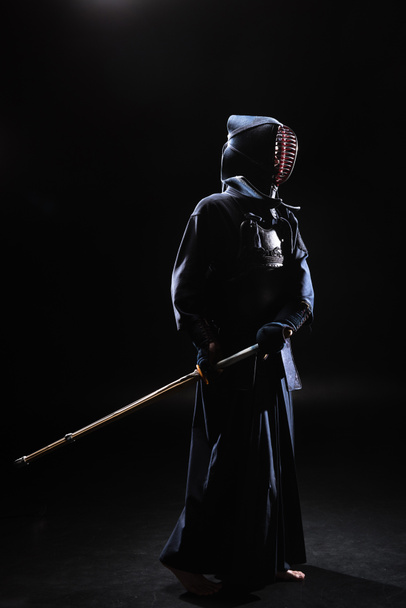 Full length view of kendo fighter in helmet holding bamboo sword on black - Фото, зображення