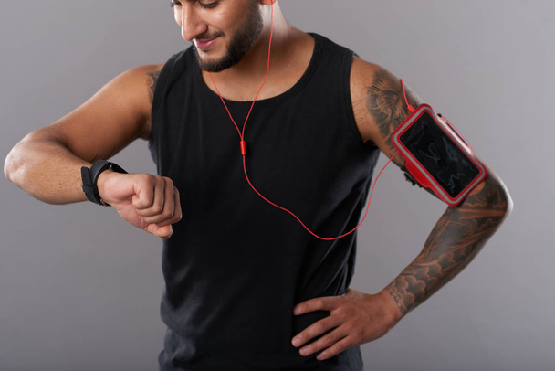 Кадрирование улыбающийся атлетичный человек с повязкой и наушниками глядя на смарт-часы во время тренировки на сером фоне
 - Фото, изображение