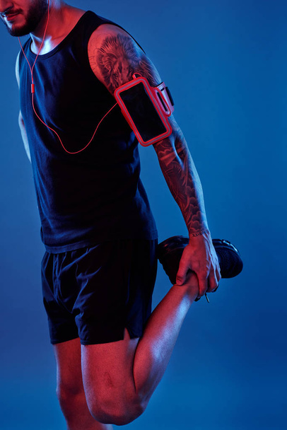 Colpo ritagliato di atletico tatuato uomo in abbigliamento sportivo e cuffie indossando bracciale con telefono e gambe flettenti su sfondo blu
 - Foto, immagini