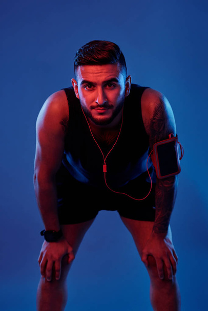 Handsome serious tattooed sportsman in armband and headphones leaning on knees and looking at camera on blue background - Fotografie, Obrázek