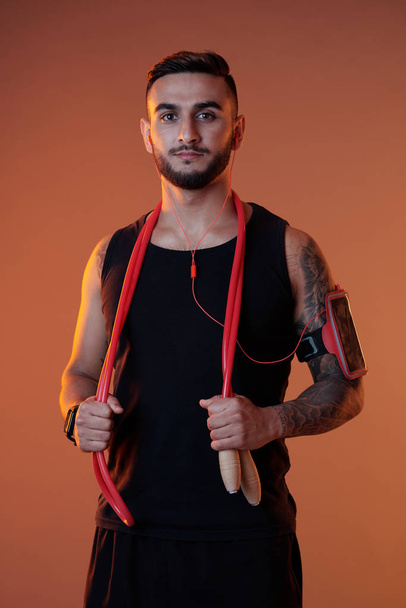 Красивый серьезный спортсмен в наушниках и повязке, держащий скакалку, глядя в камеру
  - Фото, изображение