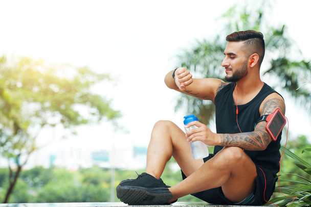 Pro dospělé vousatý muž s lahví vody nošení sluchátek a kontrola smartwatch při posezení venku - Fotografie, Obrázek