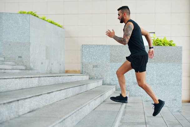 Vista lateral do homem de sportswear preto correndo escadas enquanto trabalhava ao ar livre na rua olhando focado
 - Foto, Imagem