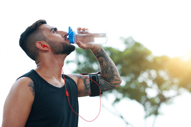 Tätowierter Sportler mit Kopfhörer und Armbinde trinkt Wasser aus Flasche auf der Straße - Foto, Bild