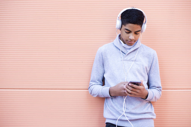 Jeune homme noir écoute de la musique sur son téléphone portable tout en se tenant dans la rue. Adolescent afro-américain dans un casque à l'aide d'un smartphone
.  - Photo, image