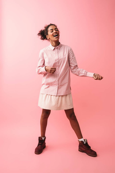 Beautiful young african woman posing isolated over pink wall background screaming singing. - 写真・画像