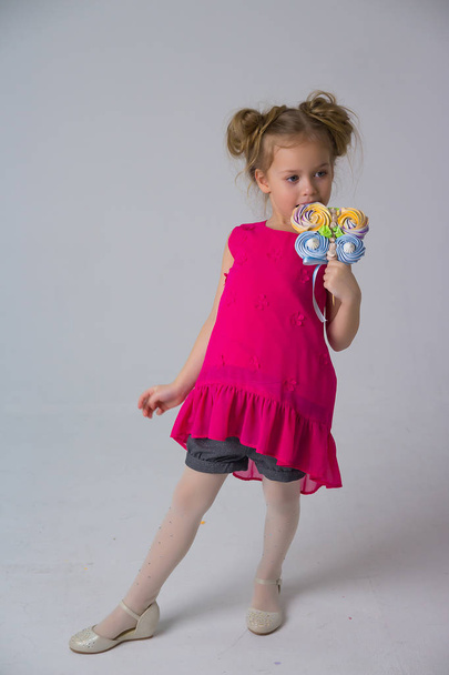 little girl in a coral dress holding a candy on a stick, bites her, sweets in childhood. sweet life. - Foto, Imagen