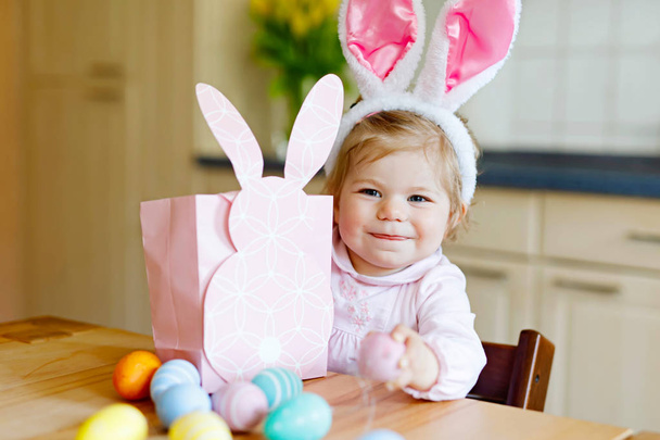 Cute little toddler girl wearing Easter bunny ears playing with colored pastel eggs. Happy baby child unpacking gifts. Adorable kid in pink clothes enjoying holiday - Photo, Image