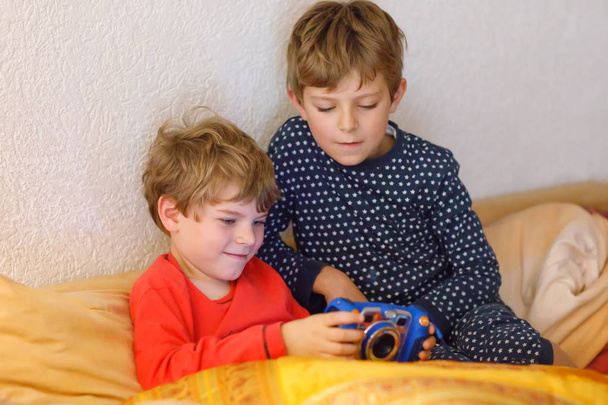 Two preschool or school kids boys, siblings and brothers having fun after school day playing video game at home and taking pictures with toy camera. Best friends, twins in pajamas or nightwear - Photo, Image