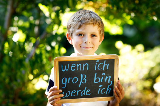 Happy little kid boy with chalk desk in hands. Healthy adorable child outdoors On desk When I grow up I want to be in German with dream profession - Foto, imagen