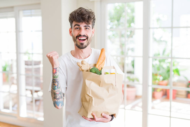 Молодий чоловік тримає паперовий пакет свіжих продуктів з супермаркету, що кричить гордо і святкує перемогу і успіх дуже схвильовані, веселі емоції
 - Фото, зображення